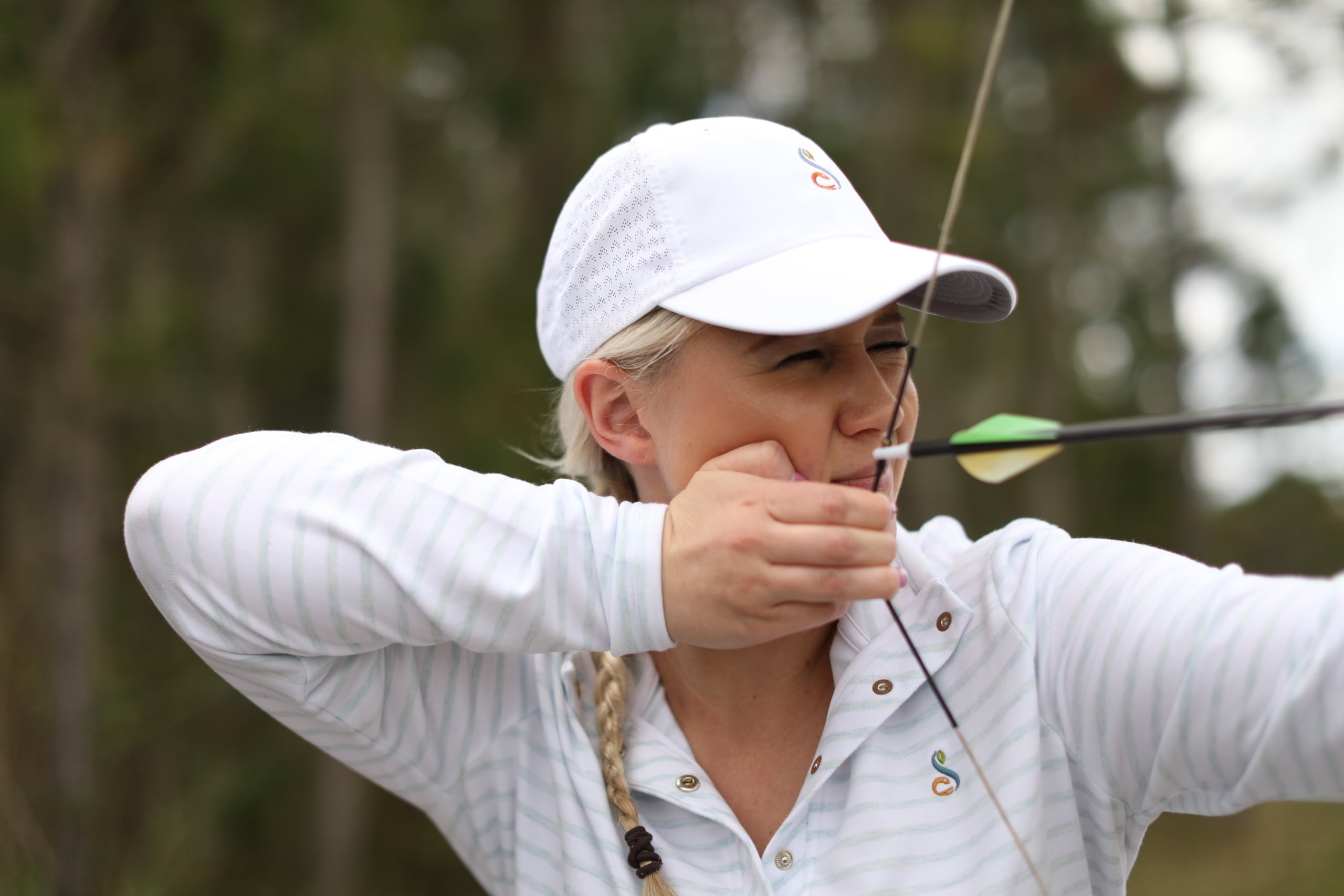 Close-up of a female archer at Streamsong taking aim with a recurve bow, wearing a white branded performance hat.
