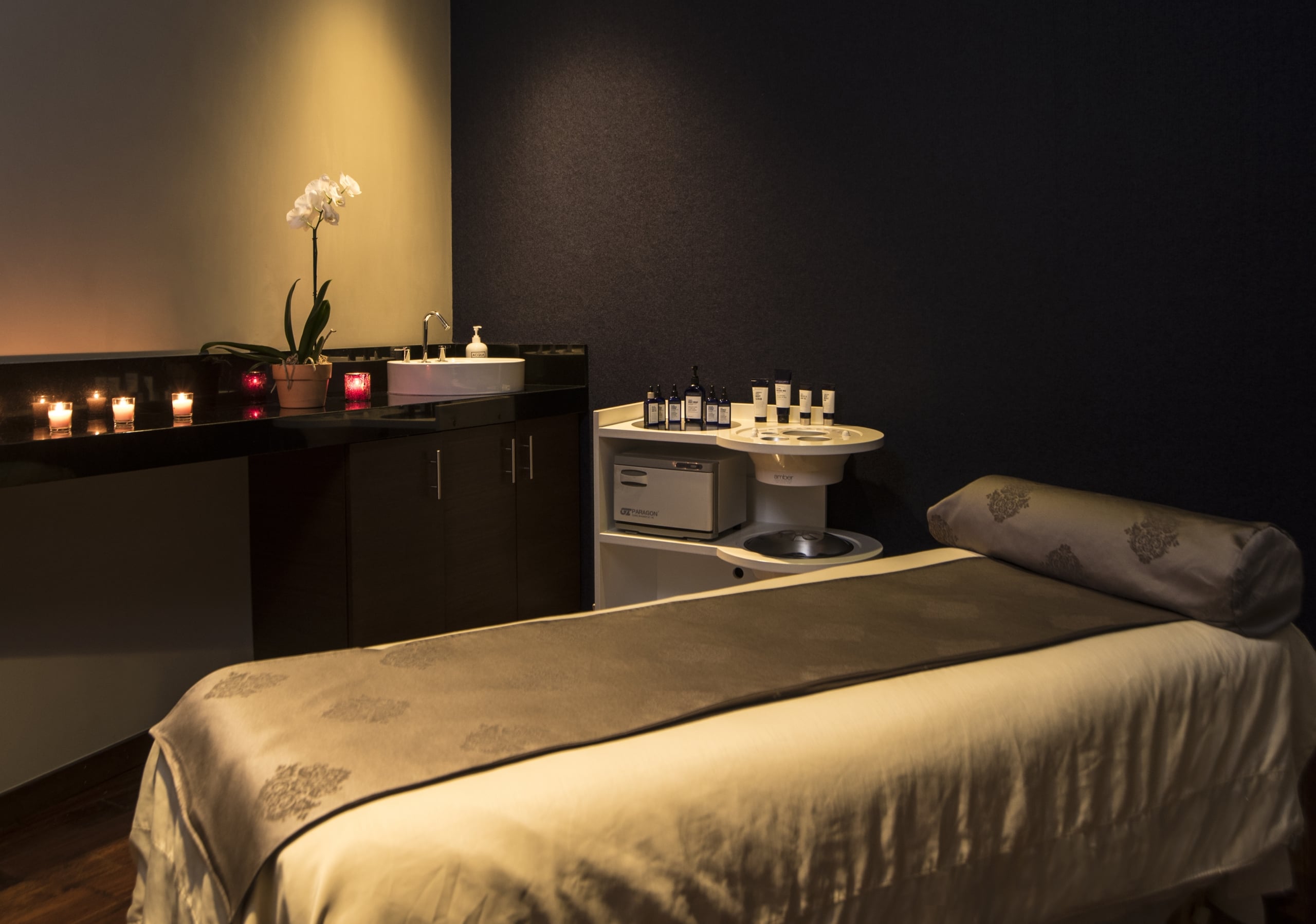 spa3-scaled.jpg Modern spa facial room featuring a sleek massage table, candlelit vanity, and high-end skincare products.