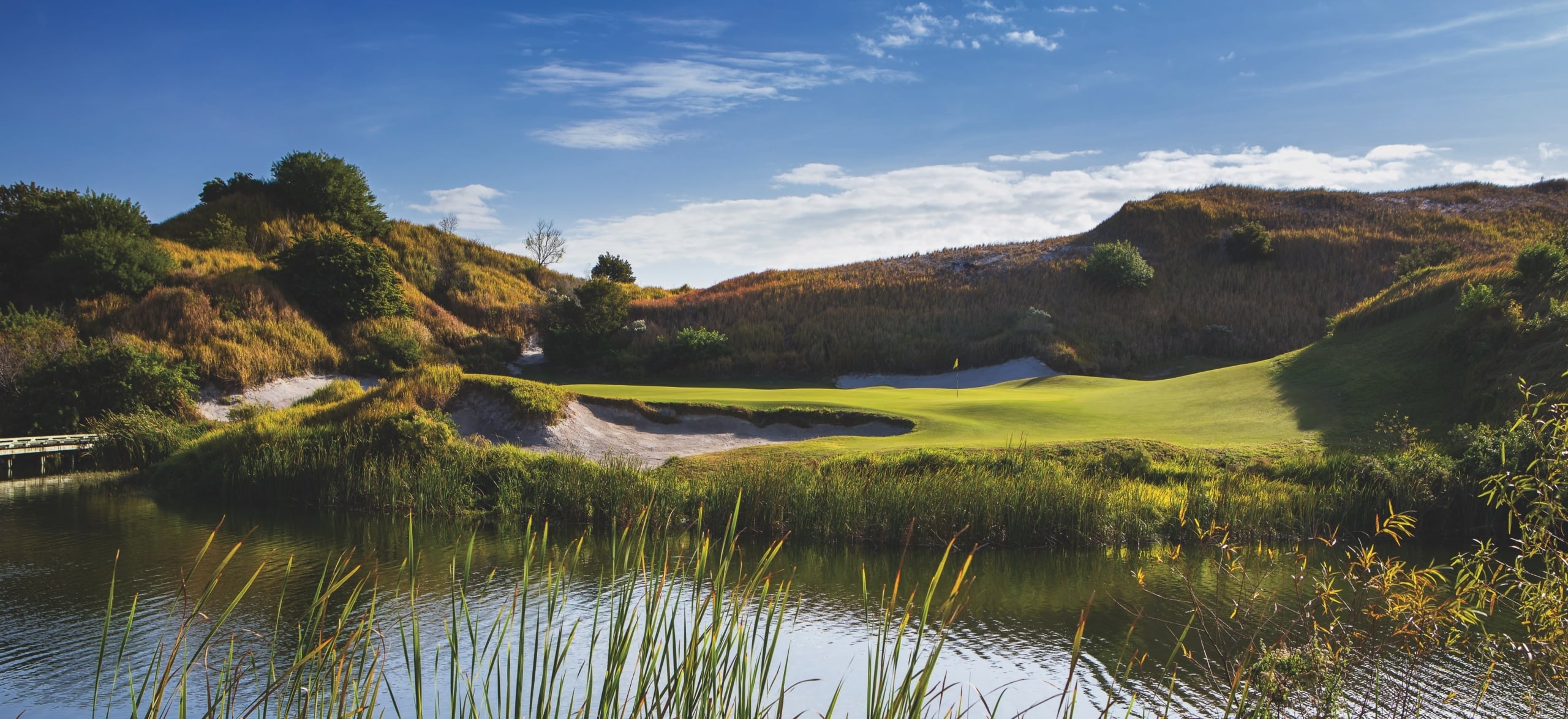 A green flanked by a bunker and a pond, with rolling hills in the background, on Streamsong Blue golf course