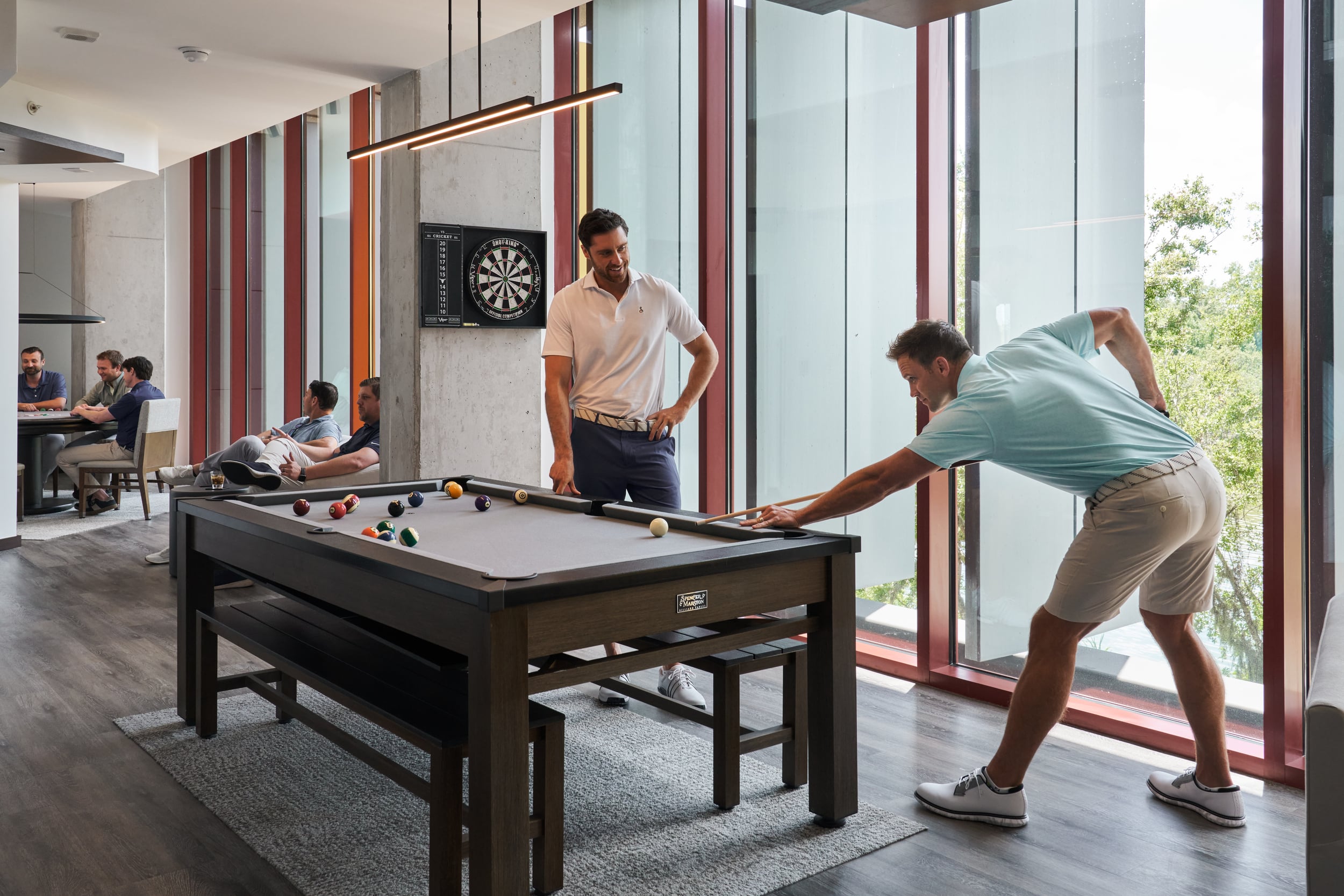 Two men play pool in front of floor to ceiling windows