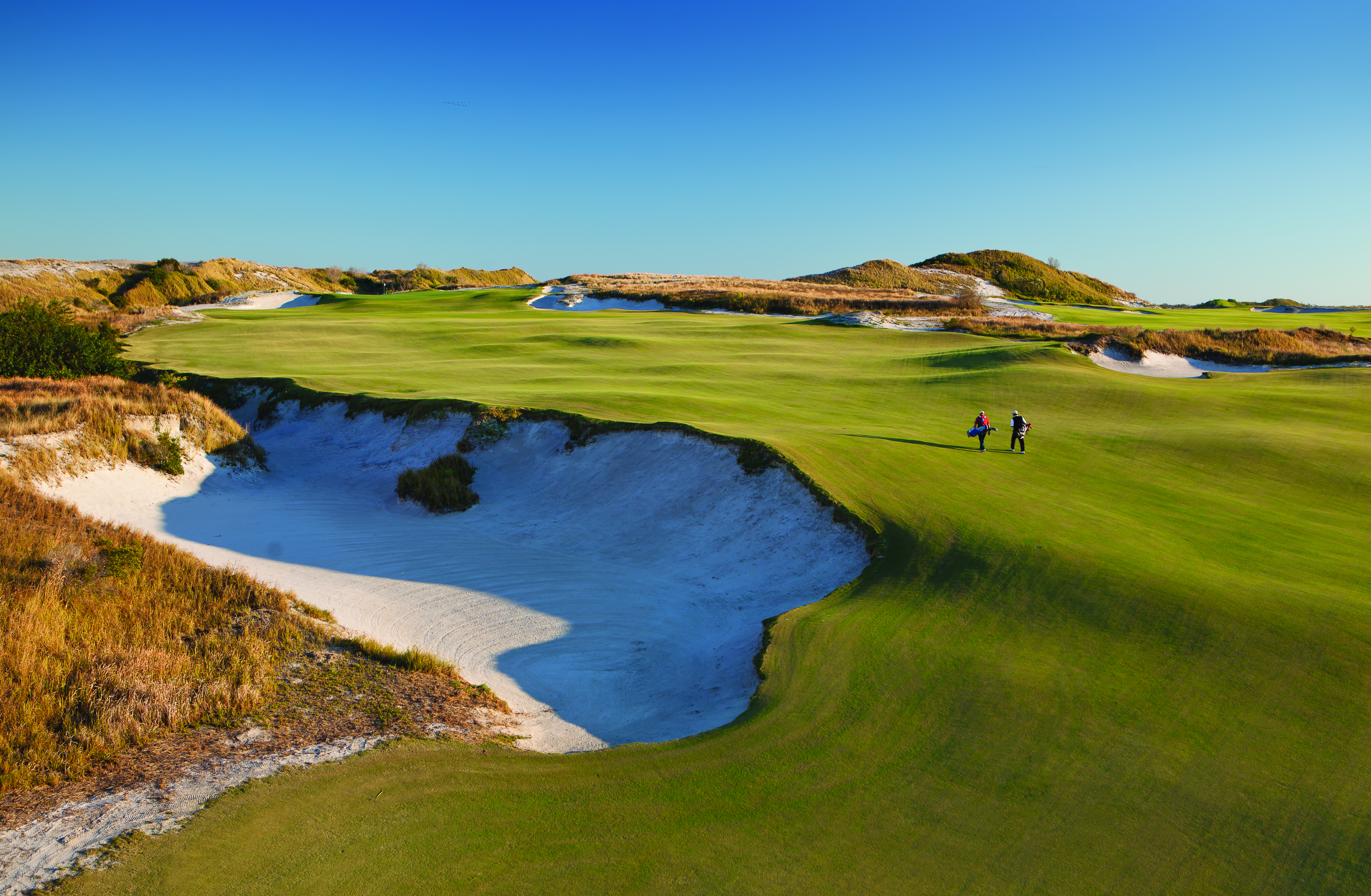Two players walk along a golf course with green grass and bunkers